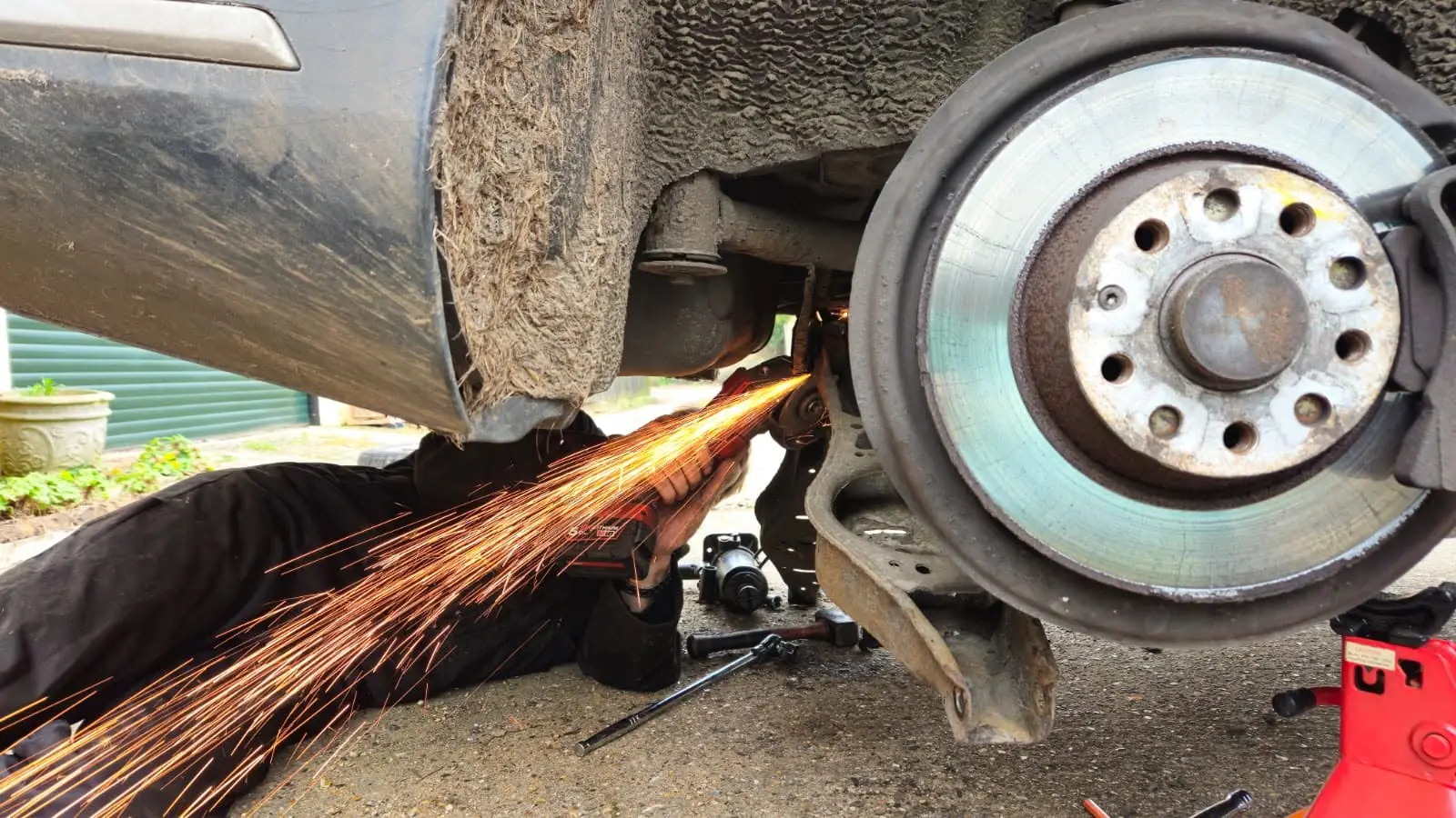 S.D.L Autos mechanic lying under a car on a driveway, cutting a seized fixing beside the rear brake disc with an angle grinder