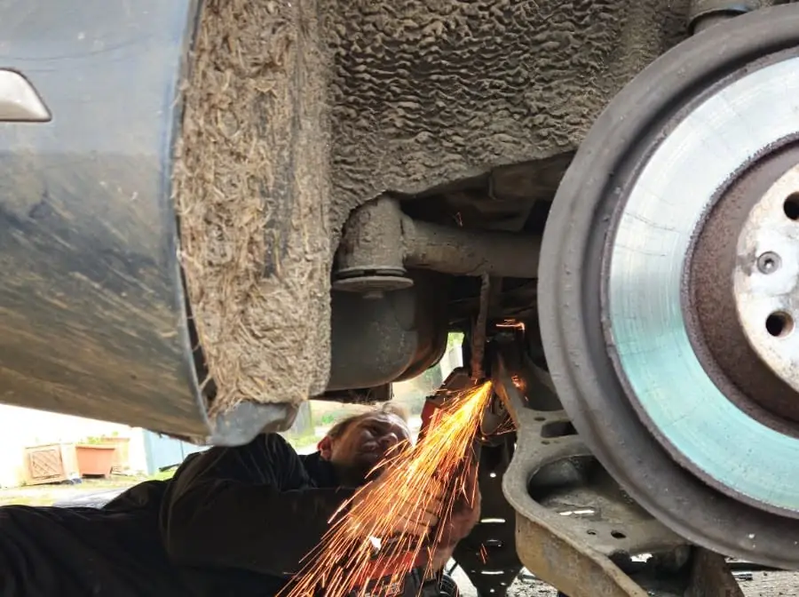 Mechanic lying under a car on a driveway, cutting a seized fixing next to the rear brake disc, with sparks flying