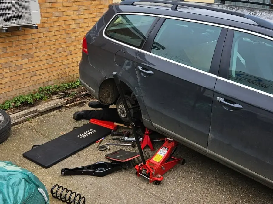 Grey VW Passat estate on a driveway with a rear wheel off, a trolley jack and an old coil spring and suspension arm on the ground