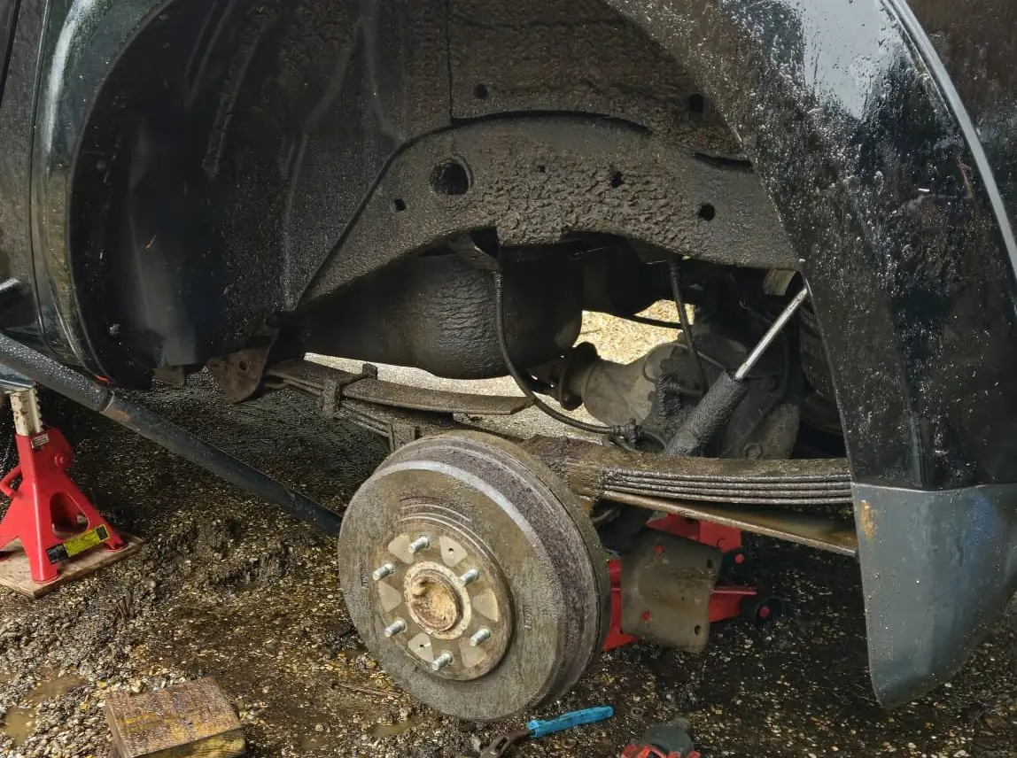 Rear wheel arch of a black pickup with the wheel off, showing the leaf spring and drum brake, with tools on the gravel