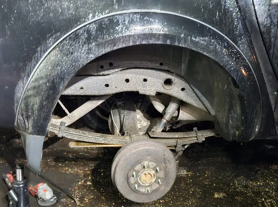Rear wheel arch of a pickup at night with the wheel removed, showing the leaf spring, drum brake and shock absorber